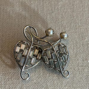 May include: Silver-toned brooch featuring two heart-shaped designs, each filled with rectangular, clear crystals. The brooch has a swirling, openwork design with two faux pearl accents.