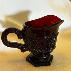 May include: A deep red glass creamer with a decorative handle and spout. The creamer has an ornate, textured design with a diamond pattern and floral accents. It sits on a hexagonal base, showcasing a vintage aesthetic.
