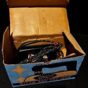 Puede incluir: Una plancha de vapor y seca Manning Bowman vintage en su caja de cartón original. La plancha negra tiene un cable en blanco y negro y un botón rojo. La caja es azul y beige con las palabras "Manning Bowman" y "Steam and Dry Iron".