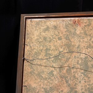 May include: A framed artwork with a wooden frame. The artwork has a mottled green and tan background. A black wire is strung across the back of the frame, secured with small nails. The frame is a dark brown color.