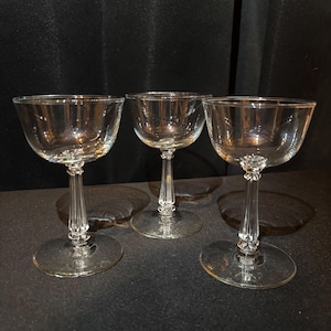 Puede incluir: Tres copas de champán transparentes. Cada copa presenta un cuenco ancho y poco profundo, un tallo decorativo y una base redonda. Las copas están dispuestas sobre una superficie oscura, con un fondo oscuro.