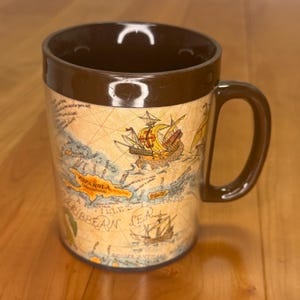 May include: A brown ceramic mug featuring a vintage-style map of the Caribbean Sea. The map displays illustrations of historical sailing ships, the text "ESPAÑOLA" and "CARIBBEAN SEA," along with various geographical labels printed in black ink.