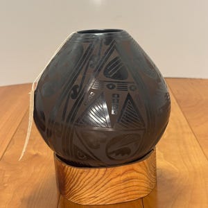 Peut inclure: Un vase rond en céramique brun foncé avec des motifs géométriques noirs, posé sur un socle en bois brun clair. Le vase présente des motifs triangulaires et courbes. Une étiquette est suspendue au vase.