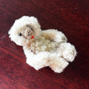 Puede incluir: Un pequeño perro de peluche vintage con pelaje color crema. El perro tiene un ojo negro, una boca roja y una mancha marrón en la espalda. El juguete está hecho de un material suave y difuso.