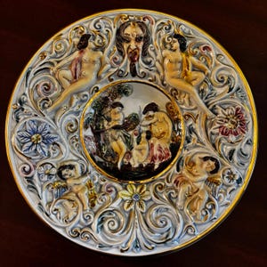 May include: A decorative ceramic plate with a detailed relief design. The plate features a central scene surrounded by figures, cherubs, and floral patterns, all in a palette of white, gold, and muted colors. The rim is trimmed with gold.