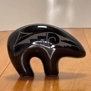 Puede incluir: Figura de oso negro brillante con un diseño estilizado en la espalda. El oso tiene un cuerpo redondeado y una forma curva de boca abierta. El diseño es de un color más claro, posiblemente gris o plateado.