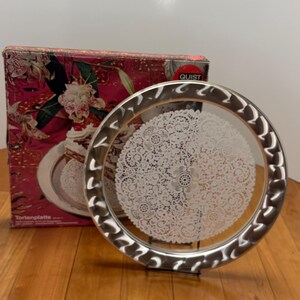 May include: A round silver-tone metal cake platter featuring a decorative embossed rim and a central white lace-patterned design. The platter is displayed in front of its original cardboard packaging, which shows a cake and the brand name Quist.