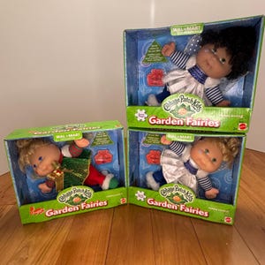 May include: Three Cabbage Patch Kids Garden Fairies dolls in their original boxes. The dolls have blonde and black hair, and are wearing fairy-themed outfits. The boxes are green and blue with the Walmart logo.