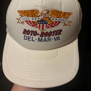 May include: Cream-colored trucker hat with a white front panel and mesh back. The front features an eagle emblem with the text "1935" and "1989", and the words "ROTO-ROOTER DEL MAR VA".