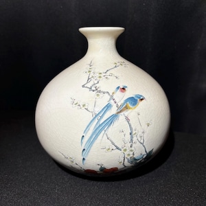 May include: Cream-colored ceramic vase with a round body and a narrow neck. The vase features a hand-painted design of two birds with blue and yellow feathers perched on a flowering branch. The background is a cracked, textured finish.