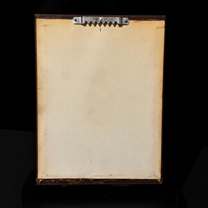May include: The reverse side of a framed picture, featuring a brown wooden frame. The backing of the artwork is a light beige hue, with a metal sawtooth hanger fixed at the top centre. The frame is dark brown.
