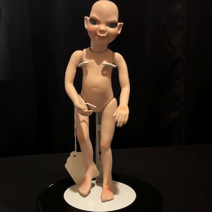 Op de afbeelding: Een huidkleurige kunstdok met een kale kop, grote oren en gedetailleerde gelaatstrekken. De pop staat op een metalen standaard met een witte basis. Het heeft beweegbare armen en benen en een label is aan de pols bevestigd.