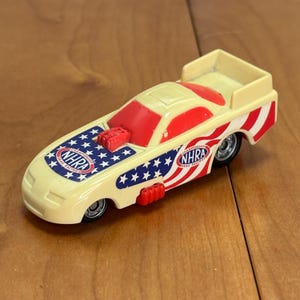 Op de afbeelding: Een speelgoed dragraceauto in cr&egrave;me, rood en blauw. De auto heeft het NHRA-logo en graphics ge&iuml;nspireerd op de Amerikaanse vlag. De auto heeft een spoiler en een gedetailleerde motor.