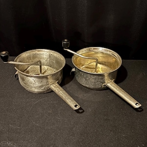 Puede incluir: Dos pasapurés vintage con cuencos y mangos de color plateado. Cada pasapurés tiene una manivela con un mango negro. Los cuencos tienen un asa lateral y un pico. Los pasapurés probablemente se utilizan para hacer puré de frutas y verduras.