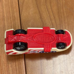 Op de afbeelding: Een rood, wit en blauw speelgoed raceauto met zwarte wielen. De onderkant van de auto heeft de tekst "NHRA Championship Drag Racing". De auto heeft een patriottisch ontwerp met sterren en strepen.