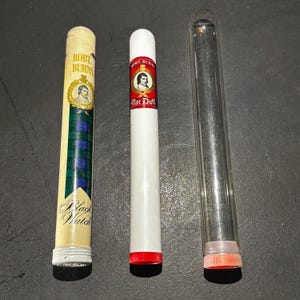 May include: Three cylindrical cigar tubes. One is off-white with a green and blue plaid design and the words "Robert Burns" and "Black Watch". The second is white with a red band and the words "Robert Burns Mac Duff". The third is clear.
