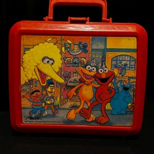 Pode incluir: Lancheira vermelha vintage com personagens da Vila Sésamo. O painel frontal mostra Big Bird, Elmo, Monstro das Bolachas e outros numa cena de rua colorida. A lancheira tem uma pega vermelha e um fecho de encaixe.