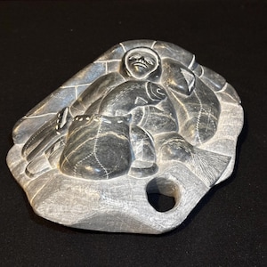 Op de afbeelding: Een grijze stenen sculptuur die een Inuit-figuur met vissen en een tas afbeeldt. De sculptuur heeft een afgeronde vorm met uitgesneden details, waaronder het gezicht van de figuur, de kleding en de schubben van de vissen. De sculptuur heeft een gat aan de onderkant.