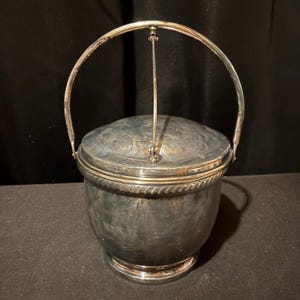 K&ouml;nnte beinhalten: Ein silberner Eisk&uuml;bel mit Klappdeckel und Henkel. Der Eimer hat einen abgerundeten K&ouml;rper mit einer dekorativen, seilartigen Verzierung am oberen Rand. Der Deckel hat ein gepr&auml;gtes Design. Der Griff ist gew&ouml;lbt und am Deckel befestigt.
