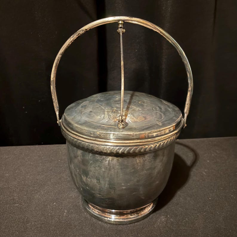 K&ouml;nnte beinhalten: Ein silberner Eisk&uuml;bel mit Klappdeckel und Henkel. Der Eimer hat einen abgerundeten K&ouml;rper mit einer dekorativen, seilartigen Verzierung am oberen Rand. Der Deckel hat ein gepr&auml;gtes Design. Der Griff ist gew&ouml;lbt und am Deckel befestigt.