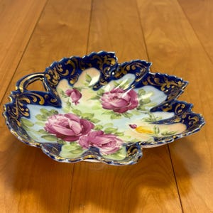 May include: A leaf-shaped decorative dish with a scalloped edge. The dish features a blue border with gold filigree and a painted floral design of pink roses and green leaves. The background is a light blue and cream color.
