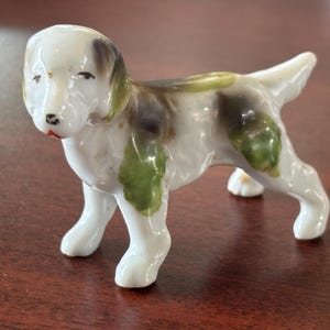 May include: A ceramic dog figurine in a standing pose. The dog is primarily white with patches of black, brown, and green. It has a red nose and black eyes. The figurine is a decorative item.