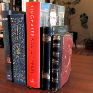 May include: A decorative bookend set in the shape of stacked books, with a black and red finish and gold accents. The bookends are holding several books with colorful covers, including a red book titled "Kingmaker Sonia Purnell."
