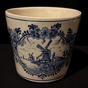 May include: Off-white ceramic planter with a blue windmill scene and floral border. The cylindrical pot features a detailed landscape design with windmills, houses, and water, and is trimmed with blue floral and scrollwork patterns.