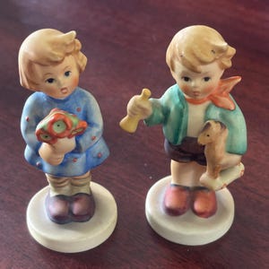 Puede incluir: Dos figuras de cerámica de niños. Una figura es una niña con un vestido azul que sostiene un ramo. La otra es un niño con una chaqueta verde que sostiene un juguete y una trompeta. Ambas figuras están sobre bases redondas de color blanquecino.