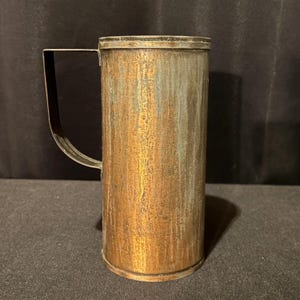 May include: A vintage copper-colored cylindrical container with a handle. The container has a tarnished, aged appearance with vertical streaks of lighter color. The handle is a dark, rectangular shape attached to the side.