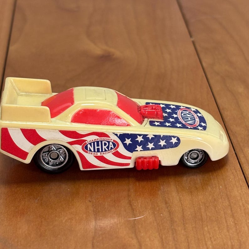 Op de afbeelding: Een speelgoed dragraceauto met een cr&egrave;mekleurige carrosserie, rode en blauwe accenten en de tekst "NHRA Drag Racing Championship". De auto heeft een patriottisch ontwerp met sterren en strepen.