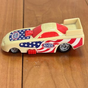 Op de afbeelding: Een speelgoed dragraceauto met een cr&egrave;mekleurige carrosserie, rode accenten en een patriottisch sterren- en strepenontwerp. De auto heeft het NHRA-logo en is een miniatuurreplica van een raceauto.