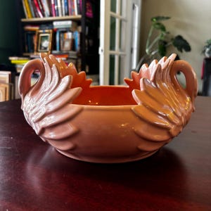 May include: A peach-coloured ceramic swan bowl with a scalloped rim. The bowl features swan heads on each side, with detailed feather designs. The interior of the bowl is a darker peach colour. The bowl is sitting on a dark wooden surface.