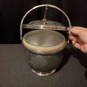 K&ouml;nnte beinhalten: Silberner Eisk&uuml;bel mit Glasinnenraum und Klappdeckel. Der Eimer hat einen Griff und ein dekoratives seilartiges Design um den Rand. Der Eimer ist aus Metall und hat eine gl&auml;nzende Oberfl&auml;che.