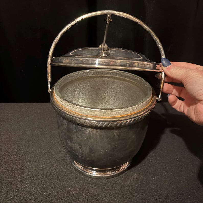 K&ouml;nnte beinhalten: Silberner Eisk&uuml;bel mit Glasinnenraum und Klappdeckel. Der Eimer hat einen Griff und ein dekoratives seilartiges Design um den Rand. Der Eimer ist aus Metall und hat eine gl&auml;nzende Oberfl&auml;che.