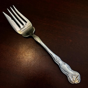 Pode incluir: Um garfo prateado com quatro dentes e um cabo decorativo. O cabo apresenta um design floral em relevo. O garfo é feito de metal brilhante e está sobre um fundo castanho escuro.
