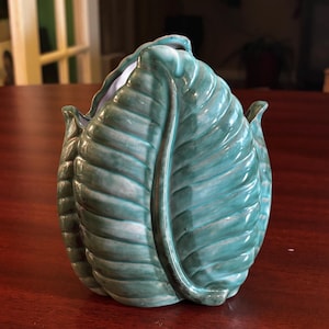 Könnte beinhalten: Ein teal-farbener Keramik-Pflanztopf in Form eines Kohlblattes. Der Pflanztopf hat eine glänzende Oberfläche und ein detailliertes Blattader-Design. Der Innenraum ist weiß. Der Pflanztopf steht auf einer dunklen Holzoberfläche.