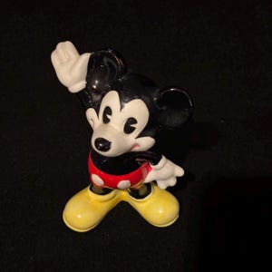 May include: A ceramic figurine of Mickey Mouse in a classic pose. The figure has a black body, white gloves, and yellow shoes. Mickey's shorts are red with white polka dots. The figurine is a collectible item.
