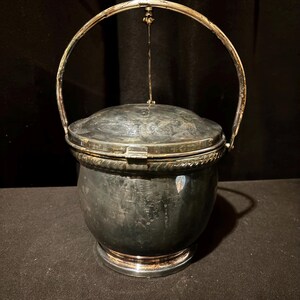 K&ouml;nnte beinhalten: Ein silberfarbener Metalleisk&uuml;bel mit Scharnierdeckel und Griff. Der Eimer hat einen runden K&ouml;rper und einen dekorativen Rand. Der Griff ist gew&ouml;lbt und am Deckel befestigt. Der Eimer steht auf einem runden Sockel.