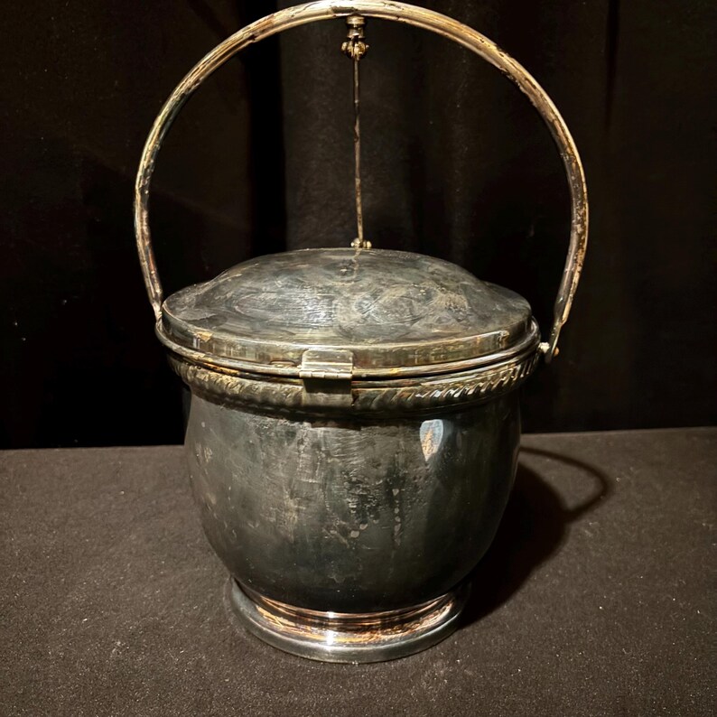 K&ouml;nnte beinhalten: Ein silberfarbener Metalleisk&uuml;bel mit Scharnierdeckel und Griff. Der Eimer hat einen runden K&ouml;rper und einen dekorativen Rand. Der Griff ist gew&ouml;lbt und am Deckel befestigt. Der Eimer steht auf einem runden Sockel.