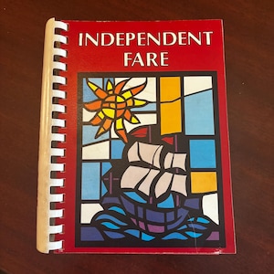 May include: A spiral-bound cookbook titled "INDEPENDENT FARE" with a red cover. The cover features a colorful illustration of a ship sailing on blue waves, a yellow sun, and a stained-glass window design.