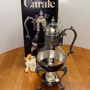 May include: A clear glass carafe with a silver-toned metal frame, ornate handle, and decorative filigree collar. The carafe features a matching silver lid and sits on a three-legged metal stand. The original black box labeled Carate is behind it.