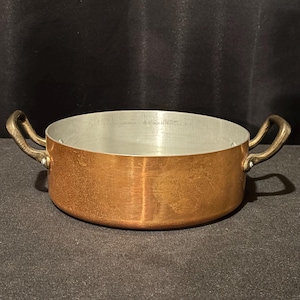 Op de afbeelding: Een koperen kookpot met twee messing handvatten. De pot heeft een zilveren binnenkant en een afgeronde bodem. De handvatten zijn gebogen en aan de zijkanten bevestigd. De pot staat op een donkere ondergrond.