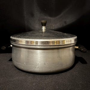 Op de afbeelding: Een vintage kookpot van geborsteld aluminium met een deksel. De pot heeft twee zwarte handvatten en een zwarte knop op het deksel. De pot is rond en heeft een zilveren afwerking. De achtergrond is zwart.