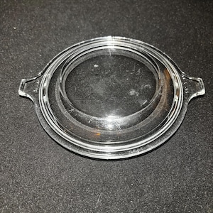 Könnte beinhalten: Ein klarer Glasdeckel mit zwei kleinen Griffen. Der Deckel hat ein kreisförmiges Design mit konzentrischen Ringen und ist wahrscheinlich für eine Auflaufform oder ähnliches Kochgeschirr geeignet. Das Glas ist transparent und ermöglicht die Sichtbarkeit des Inhalts.