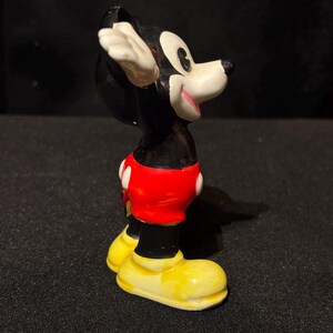 May include: A vintage Mickey Mouse figurine. The figure is primarily black with white gloves and ears, red shorts with white buttons, and yellow shoes. The figurine is posed with one arm raised and a smiling expression.