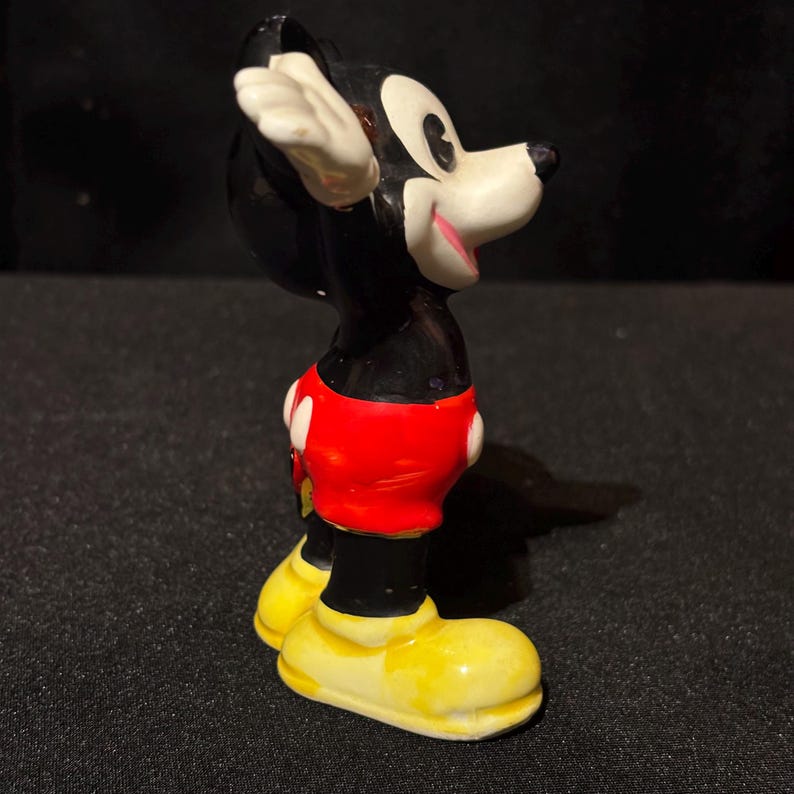 May include: A vintage Mickey Mouse figurine. The figure is primarily black with white gloves and ears, red shorts with white buttons, and yellow shoes. The figurine is posed with one arm raised and a smiling expression.
