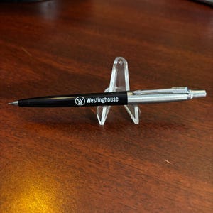 Puede incluir: Bolígrafo negro y plateado con el texto "Westinghouse" en el cuerpo. El bolígrafo se exhibe sobre un soporte de plástico transparente, sobre una superficie de madera oscura.