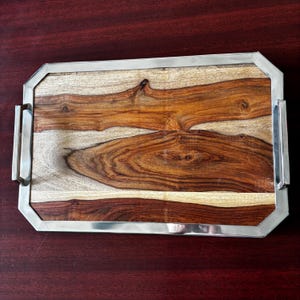 Może przedstawiać: Prostokątna drewniana taca do serwowania z polerowaną srebrną metalową ramą i uchwytami. Powierzchnia tacy ma naturalny wzór słojów drewna w odcieniach brązu, beżu i kremu. Przeznaczona do serwowania potraw lub eksponowania przedmiotów dekoracyjnych.