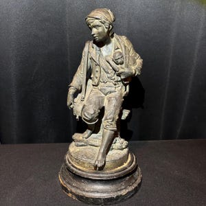 May include: Bronze statue of a seated figure in period clothing, holding a small object and a satchel. The statue is mounted on a dark, round base. The figure is wearing a hat and appears to be looking down. The statue has a weathered appearance.